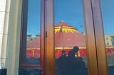 In a reflection of an unmarked storefront is a gray silhouette of a man using an electric wheelchair. Behind the man is a spectacular red and yellow circus tent.