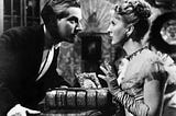 A man and a woman in Victorian evening dress looking at each other across a table. The man appears to be challenging the woman. A large gilt embossed bible sits between them.