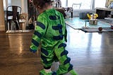 A toddler in a green dinosaur costume facing away from the camera.