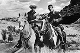 Black-and-white photo of the Lone Ranger, a man wearing a mask covering his eyes and a cowboy hat, sitting on a white horse (left) and Tonto, a Native American, sitting on a multi-color horse (right) set against a rugged background.