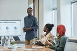 Data Analyst standing next to a screen with pie charts, presenting in a conference room with two people seated at the table.