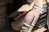A ballerina, dressed in a dark leotard, with sheer white stockings. She wears off-white leg warmers, just over her ankle and the heel of her ballet shoes. She sits in a corner, with stacks of books surrounding her, filling the frame. We can’t see the spines of the books, just the pages. They’re all thick, holding, eluding to millions of words. Her head is buried deep in a huge tome as her hands cradle the book gently, dark brown hair obscuring her face. A white lamp illuminates the scene.