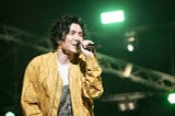 Fujii Kaze in a yellow jacket, singing into a mic, with a green light in the background.