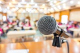 Close-up of a microphone on a stage, with a softly blurred backdrop of an audience in a brightly lit auditorium.