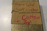 Shiny gold jewelry box, on the lid of which are two pieces of masking tape with these words in red ink: “Laura Lind look under Cotten in Box”