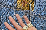 Prison, barbed wire, chain link fence, captive, yellow flag, in the foreground is an open hand holding Scrabble tiles which spell out the word Help