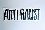 Hand lettering of “Anti-racist” in all capitalized letters painted in black ink on white strip of paper.