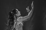 Black and white photo of a young woman with long, flowing hair in a dance pose, her arms elegantly raised, expressing intense emotion.