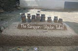 A traditional cooking setup from a tribal village with “The Sangyan: Ulgulan Talks!” written over it on the smaller platform and below the earthen chimney. The setup has eight small earthen chimneys over a small platform that settles over a larger platform all made up of soil and other natural ingredients and are brown in colour.