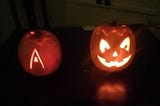 Two pumpkins, one a traditional jack-o-lantern, the other with the letter A carved into the front, both lit up from inside against a dark background