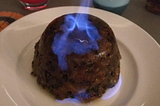 A Christmas pudding sits on a white plate with a blue flame rising out of the top.