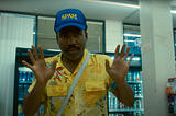 A Black man in a floral shirt and a “Spam” hat has his hands up