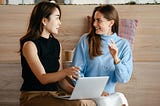 Two women talking to each other about work with a laptop in front of wooden stadium seating