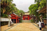 The bright red entrance of a traditional Japanese Shinto Shrine contrasts with the lush greenery surrounding it.