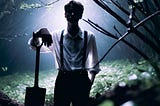 A young man in silhouette leans on a shovel in a dark glade