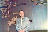 Photo of a smiling middle age man in a suit leaning against a wall chart of global financial reporting.