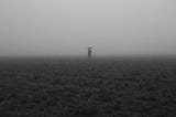A black & white photo with a person standing on empty land, holding umbrellas, vanishing into the mist.