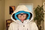 A woman bundled up in a down jacket and a hoodie frowns at the camera.