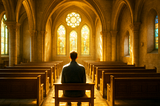 Image of a solitary man sitting on a wooden pew inside a bright, sunlit church, facing the stained-glass windows at the front, with soft golden light filling the space and highlighting the empty rows of pews around him.
