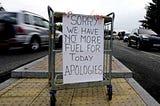 A white sheet of card tied crudely to a metal trolley on a yellow pedestrian crossing that reads “SORRY WE HAVE “NO” MORE FUEL FOR TODAY APOLOGIES” written by hand in capital letters