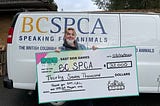 woman holding a large novelty cheque made out to the BC SPCA from East Side Games with a value of $37,000 while standing in front of a white van with BC SPCA branding.