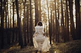 A woman lifts up her white skirt while walking through a forest.