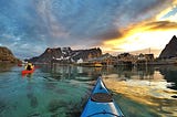 The view from my blue kayak. Craig is in a red kayak in front of me and there are steep mountains ahead. The sea is turquoise in areas and the grey and orange clouds are reflected in the water.