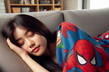 A young woman is sleeping on a couch. She has long straight dark hair. There is a blanket pulled up over her shoulders with an image of Spiderman on it.