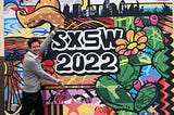 The author poses smiling in front of a colourful sign advertising SXSW 2022, wearing a grey jumper and black trousers.