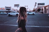 A girl is walking in a city covering her face with her blond hair, few cars are passing on the road