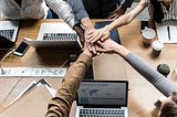 A group puts hands in the middle of the table containing several computers. Crowdsourcing.