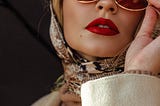 Haughty young women with deep red lipstick and scarf covering her head with outdoor jacket on peering down from her designer sunglasses