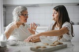 Woman with gray hair speaking to young girl with cookie in her mouth at kitchen table
