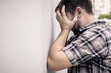 A frustrated man holds his head in his hands and leans against a wall.