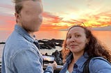 Beautiful colors of a sunset with the sky in the upper half of the photo and water in the lower half with the woman on the right smiling while looking into the camera and the man on the left with his face blurred out.