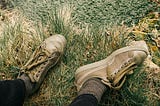 Top-down view of a person’s feet in worn, olive-green sneakers with loose laces, resting in patchy grass near a stone edge. The person’s legs are relaxed and slightly apart, as if pausing or observing from the sidelines.