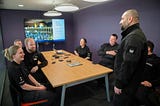 Six MDP officers, men and women, gathered in a conference room, sitting around a table, listening to PS Rahat who is standing and presenting about Ramadan. In the background there is a large digital screen with the image of a slide on it from PS Rahat’s presentation.