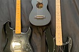 The photo shows three musical instruments: an electric guitar, a guitalele (super small classic guitar), and a bass guitar.