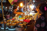 night market outdoor food stall in Siem Reap with woman serving from many bowls of food while customers look on