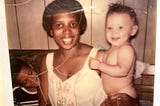 A Black woman smiling, looking straight into the camera while sitting on a couch with a pale skin baby in her left arm and a boy with a darker complexion sitting on her right, smiling and looking over at the baby.