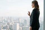 A high-achieving leader — gender neutral presentation, early-to-mid 40s, sharp professional dress — standing at a floor-to-ceiling window in a high-rise office. City skyline visible below. Everything they have built is literally beneath them. The posture is upright. The expression is not triumph — it is absence. The surface is intact. The fracture is not visible. Lighting: cool, natural, slightly overexposed. No text overlay. The image communicates: everything has been built. Something is missin