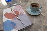 Colourful notebook with a pen and a blue cup of espresso on a marble countertop