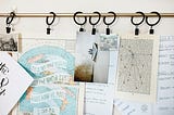 A collection of papers, maps, and images hangs from black clips on a gold curtain rod, creating a writer’s mood board. The papers include handwritten notes on a world map, a page of typed text with annotations, and a whiteboard with notes on story structure.