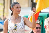 A Middle-Aged Woman With A Ponytail Wearing A Polka Dot Top Taking A Selfie In A Public Space.