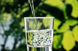 Close-up of water being poured into a clear glass, with bubbles forming as the glass fills. Green, blurred leaves are visible in the background, suggesting an outdoor setting.