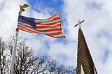 A U.S. flag hangs on an eagle-topped flagpole against a blue sky. To its right is a church steeple, nearly equal in height.