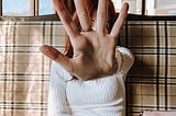 Woman with long read hair sitting on a plaid covered sofa. She is wearing a white ribbed sweater. Her left arm is out-strecthed and her hand is raised with palm facing the reader in the center of the photo.