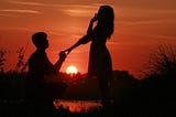 A young man proposes to a young woman, silhouetted by a red sunset