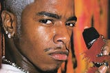 An image of rapper, Sisqó, staring at the camera with a smirk on his face. His hair is short and died white. He has diamond earrings and a silver chain around his neck. He’s holding a microphone in one hand.