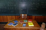 Image of books spread across a teacher’s desk in front of a classroom chalkboard.